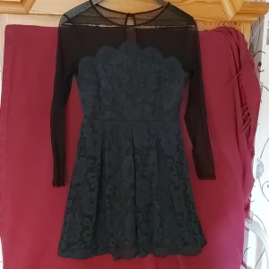 Svart spetsklänning med lång ärm - En elegant grön spetsklänning med lång ärm och transparenta detaljer vid axlar, armar och hals. Klänningen har ett vackert mönster och en figurnära passform med en något utsvängd kjol. liten i storkek