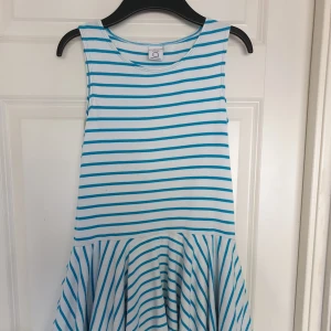 Randig klänning från Polarn O. Pyret - En vit och blå randig klänning från Polarn O. Pyret. Klänningen är ärmlös och har en vid kjol med volanger. Tillverkad i Litauen.