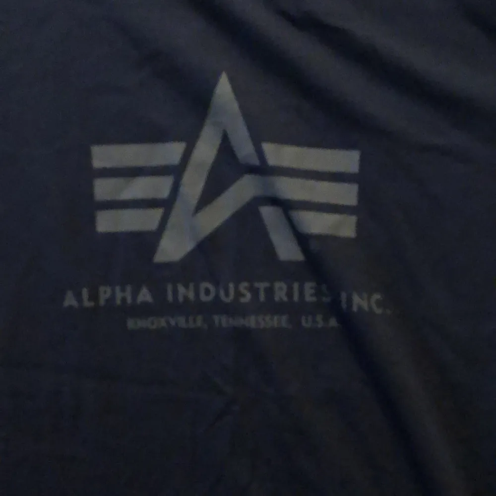 En mörkblå t-shirt från Alpha Industries med deras logotyp tryckt på framsidan. T-shirten har korta ärmar och en rund halsringning.. T-shirts.