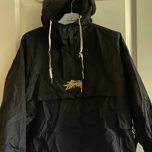 Svart vindjacka från Stüssy - En svart vindjacka från Stüssy med en stor gul logotyp på framsidan och baksidan. Jackan har en huva med vita dragsnören och en halv dragkedja framtill. Den har även en stor ficka på framsidan.