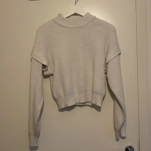 Beige stickad tröja - En vit stickad tröja med långa ärmar och ribbad struktur. Tröjan har en unik design med extra tygdetaljer över axlarna. Den är varm och bekväm, perfekt för sena sommarkvällar