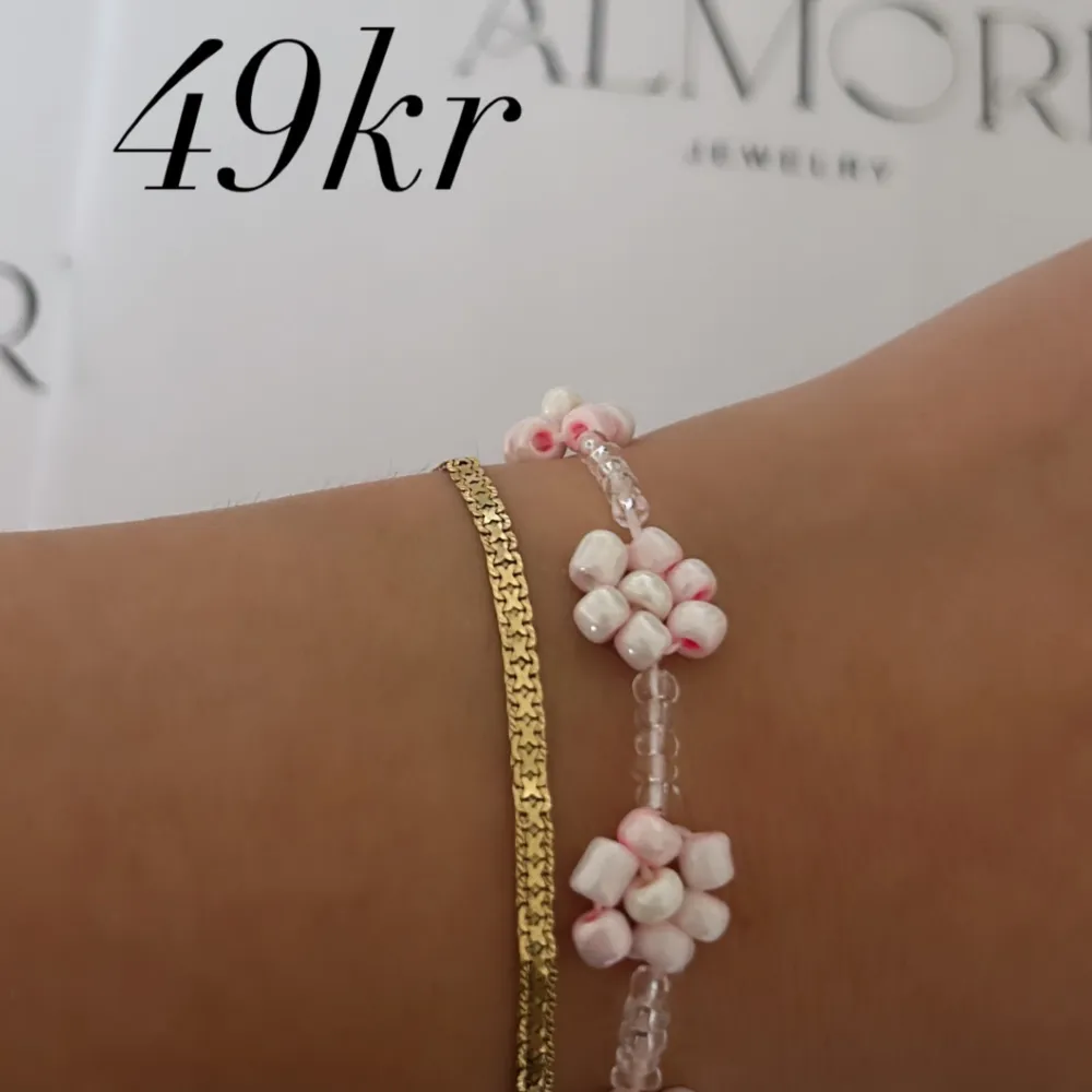 Ett vackert armband med små vita och rosa blommor samt gulddetaljer. Armbandet har en elegant och feminin design.. Accessoarer.