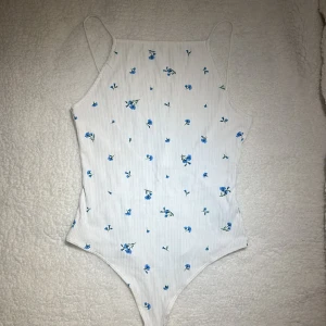 Blommig och vit bodysuit  - Sommrig vit bodysuit med blå blommor på. Väldigt bra skick och har inte använts mer än ett par gånger och aldrig utan leggings under. Tvättas i 30 grader och är i storlek M. 