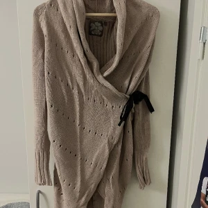 Ribbstickad kofta - Ljusbrun ribbstickad omlottkofta. Sparsamt använd. Håller värme och är väldigt mysig. Knytband precis i höjd med bysten.