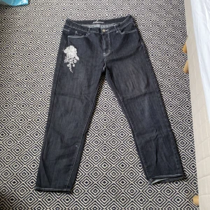 Svarta jeans med vit blomma - De här är i bra skick och har ett snyggt broderi på. Midjevidden är 42 cm och inner benslängden är 65 cm. 