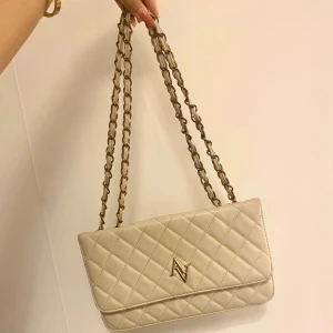 Beige quiltad axelväska - Säljer en snygg beige axelväska med quiltat mönster. Väskan har en kedjerem och ett elegant guldmärke på framsidan. Perfekt för både vardag och fest! 👜✨