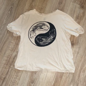 Beige t-shirt med fisktryck från H&M Divided - Säljer en begie t-shirt från H&M Divided i storlek S. T-shirten har ett coolt tryck med två fiskar i en yin-yang design på framsidan. Den är kortärmad och gjord av bomull, vilket gör den superbekväm att ha på sig. Perfekt för en avslappnad stil!🪷☯️
