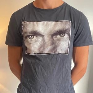 Svart t-shirt med ögonmotiv - Säljer en svart t-shirt med ett coolt ögonmotiv på bröstet. T-shirten är i normal passform och har korta ärmar. Perfekt för en avslappnad stil eller för att sticka ut på festen. Materialet känns som bomull och är bekvämt att ha på sig. Passar bra till jeans eller shorts!