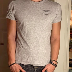 Aston Martin Racing Grå t-shirt - En grå t-shirt från Aston Martin Racing med korta ärmar och en bra passform. T-shirten har en liten logotyp på bröstet. Storlek M men passar som S. Modellen är 182 cm och väger 75 kg. Pris går att diskuteras!