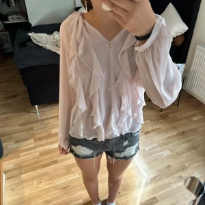 Ljusrosa blus med volanger - En elegant ljusrosa blus med volanger framtill och långa ärmar. Blusen har en lös passform och är tillverkad i ett lätt och luftigt material.