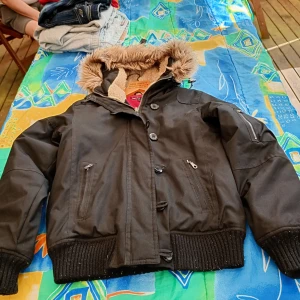 Svart lumberjacka fodrad luva stl 34/36 - Fin lumberjacka med luva och fodrad o resår i midjan o ärmarna samt knäppes med dragkedja och knappar,fickor fram mm.viss slitage förekommer se bilder.