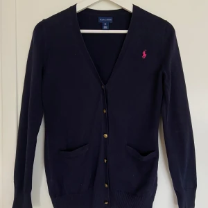 V-ringad bomullskofta med guldknappar från Ralph Lauren Kids - XL (16 år) - Ursöt och tidlös kofta från Ralph Lauren kids i 100% bomull. Koftan har en klassisk v-ringning, guldknappar och två små fickor på framsidan. Storlek: XL - 16 år, men kan säkert passa en XS dam också. Slutsåld.