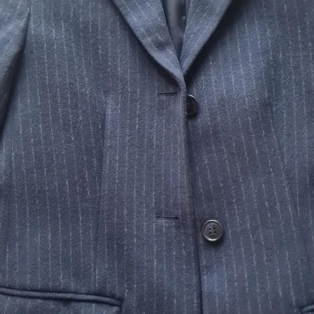 Marinblå kritstrecksrandig kappa. Mycket gott skick. Material: Yttre- 90% ull & 10% kashmir Innre- lika delar polyester & viskos. Takit.