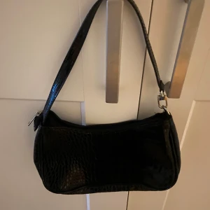 Svart handväska - svart handväska med en glansig yta och krokodilmönster. Väskan har en dragkedja upptill och en axelrem för enkel bärning. Mycket rymlig💕