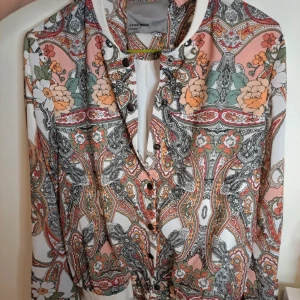 Mönstrad jacka från Vero Moda - En färgglad mönstrad jacka från Vero Moda med blommor och paisley-mönster i nyanser av rosa, grön, orange och vit. Jackan har långa ärmar och knappar framtill.