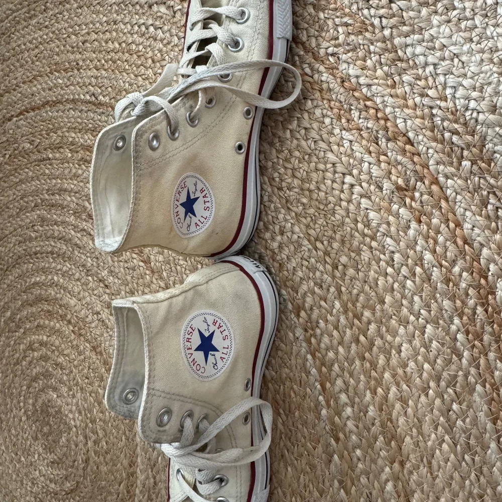 Ett par beige höga sneakers från Converse med snörning och vit sula. Skorna har den klassiska Converse-logotypen på sidan och en röd rand längs sulan.. Skor.