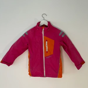Rosa orange skal jacka barn - Funkar perfekt nu när det börjar bli lite varmare då det är en skal jacka. Är i storlek 122 och är andvänd men i bra skick. Passar både tjej och kille. Saknar Luva.