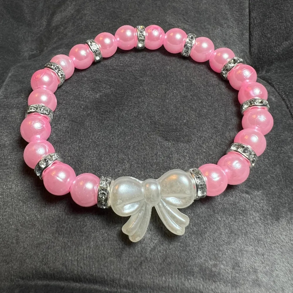 Pärlarmband med rosa pärlor, blingiga mellanplattor och en söt vit rosett.🎀 Är gjord med gummitråd och är ca 17 cm lång.. Accessoarer.
