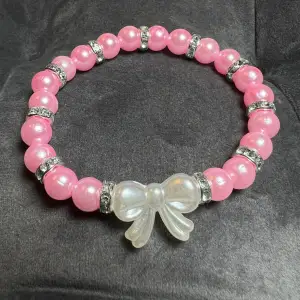 Pärlarmband med rosa pärlor, blingiga mellanplattor och en söt vit rosett.🎀 Är gjord med gummitråd och är ca 17 cm lång.