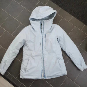 Helly Hansen skidjacka - Skidjacka ifrån Helly Hansen! Köpt förra året så enbart använd en säsong! Fodrad med dun på insidan så den håller värmen bra! Tyvärr har den fått ett litet jack på högra ärmen (bild 3) men skickar med laglapp i samma blåfärg, så lätt att laga!