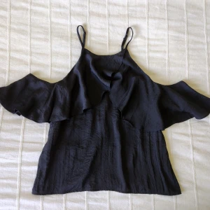 Svart topp - volangtopp - satin - storlek 34 - Svart volangtopp i ett satinliknande lite glansigt material 🖤 Knäppning i ryggen och med reglerbara axelband. 