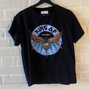 En svart t-shirt från Sisters Point med ett tryck av en örn och texten 'King of Wild Tour' i blått och grönt. T-shirten har korta ärmar och en rund halsringning.