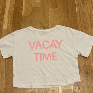 Vit t-shirt med texten 'VACAY TIME' från Cool Club - En vit t-shirt från Cool Club med texten 'VACAY TIME' i rosa på framsidan. T-shirten har korta ärmar, en rund halsringning och den är litr croppad.