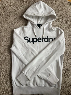 Vit hoodie från Superdry - Säljer en stilren vit hoodie från Superdry med svart logga över bröstet. Den är i gott skick men kan vara lite nopprig på vissa ställen såsom på muddarna och i luvan, den har även fått en missfärgning precis vid ändan av höger snodd (se bild 4) men det är inget som märks. Nypris: 800kr