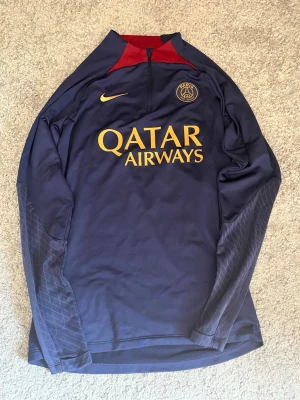PSG tröja - Snygg mörkblå långärmad träningströja från PSG med gul Nike-logga och Qatar Airways-tryck på bröstet. Tröjan har halv dragkedja, röd detalj vid kragen och diskret mönster på ärmarna. Perfekt för fotbollsträning eller chill.