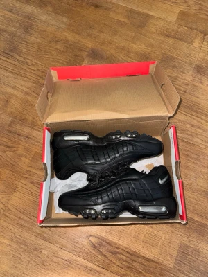 Nike Air Max 95 svarta sneakers - Säljer ett par svarta Nike Air Max 95 sneakers med synliga Air-enheter i sulan och reflekterande detaljer. Skorna har en cool, sportig look med svarta skosnören och en mix av läder och mesh-material. Perfekta för dig som gillar streetwear och klassisk Nike-stil.