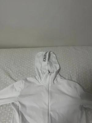Vit hoodie med dragkedja och tryck - Snygg vit hoodie med dragkedja och huva. På huvan finns ett svart tryck med text och på bröstet ett litet svart motiv. Perfekt för en chill och stilren look. Mjuk bomullskänsla och klassisk passform. Anpassad även för sport. 