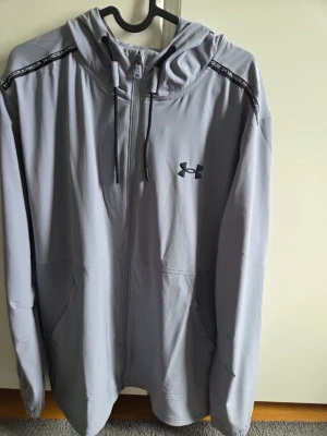Grå zip hoodie från Under Armour - Snygg grå zip hoodie från Under Armour med svart logga på bröstet och textdetaljer på axlarna. Tröjan har huva med dragsko, fickor på sidorna och är tillverkad i ett mjukt funktionsmaterial. Perfekt för träning eller chill.