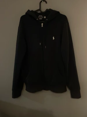 Svart hoodie med dragkedja och huva - Säljer en blå hoodie med dragkedja och huva från polo Ralph lauren. Den har två fickor framtill och en liten vit logga broderad på bröstet. Hoodien har långa ärmar och en klassisk, avslappnad passform. Perfekt för chill dagar eller när du vill ha en enkel och stilren look.