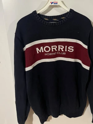 Morris Rowing Club stickad tröja - Mörkblå stickad tröja från Morris med bred vinröd och vit rand över bröstet och texten 'Morris Rowing Club' broderad framtill. Tröjan har rund halsringning och ribbade muddar vid ärmslut och nederkant. Perfekt för en sportig och stilren look. Storlek Xl men passar perfekt på L. Pris kan diskuteras vid snabb affär.