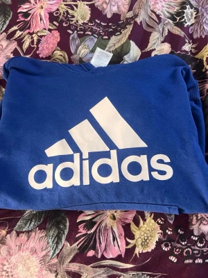 Blå hoodie från Adidas med logga - Snygg blå hoodie från Adidas med stor vit logga och text på bröstet. Klassisk modell med huva och ribbade muddar. Perfekt för en sportig och avslappnad stil.