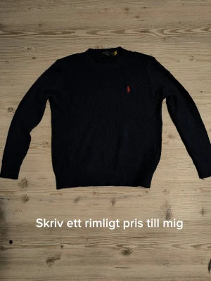 Oanvänd äkta stickad Ralph Lauren - Klassisk mörkblå kabelstickad tröja från Polo Ralph Lauren med rund halsringning och den ikoniska röda loggan broderad på bröstet. Tröjan har ribbade muddar vid ärmslut och nederkant. Perfekt för en stilren och tidlös look.