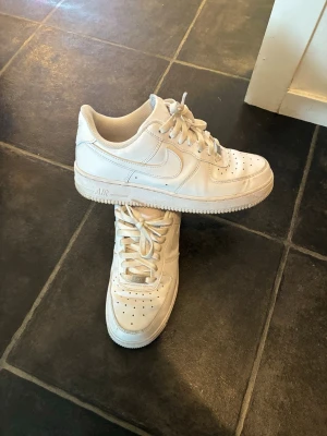 Vita Nike Air Force 1 sneakers - Klassiska vita Nike Air Force 1 sneakers i lågt utförande. Skorna har en chunky sula, perforerad tåbox och ikonisk Swoosh på sidan. Tillverkade i läder med vita skosnören och detaljer. Perfekta för en clean och stilren look.