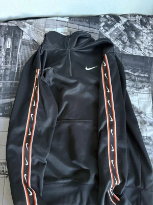 Svart Nike hoodie med dragkedja - Säljer en svart hoodie från Nike med halv dragkedja framtill och vita Nike-loggor längs båda ärmarna. Hoodien har röda och vita ränder på ärmarna och en hög krage. Perfekt för en sportig och avslappnad stil.
