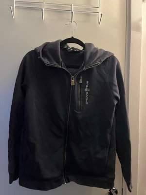Svart hoodie från Sail Racing - Snygg svart hoodie från Sail Racing med dragkedja framtill och logga på bröstet. Hoodien har en stor huva och två fickor på sidorna. Tillverkad i mjukt material som känns skönt mot huden. Perfekt för dig som gillar en clean och sportig stil.
