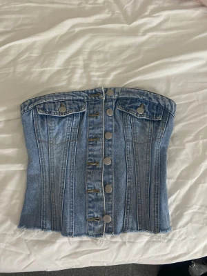 Jeanstopp - Snygg denim korsett-topp i ljusblått med knappar framtill och två dekorativa fickor. Toppen är axelbandslös och har råa kanter nertill samt smock baktill för en bekväm passform. Perfekt för en trendig och edgy look.