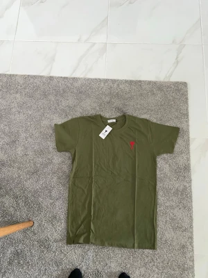 Olivgrön t-shirt från Ami - Snygg olivgrön t-shirt från Ami med klassisk passform. T-shirten har korta ärmar och ett rött broderat hjärta med bokstaven A på bröstet. Tillverkad i mjuk bomull som känns skön mot huden. Perfekt till jeans eller shorts.