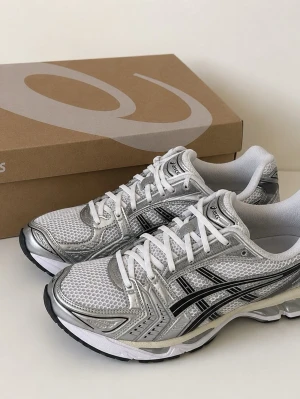Asics gel kayano 14 - Snygga sneakers från Asics i silver och vitt med svarta detaljer. Ovandelen är i mesh och syntetmaterial för bra andningsförmåga. Skorna har en sportig look med klassisk snörning och dämpad sula. Perfekta för dig som gillar en modern och clean stil.
