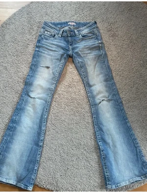 Botcut jeans från Gina Young  - Säljer ett par blå bootcut jeans med slitningar på låren och knäna. Jeansen har klassisk femficksmodell, låg midja och ljus tvätt med snygga detaljer. Perfekta för en avslappnad och trendig look.