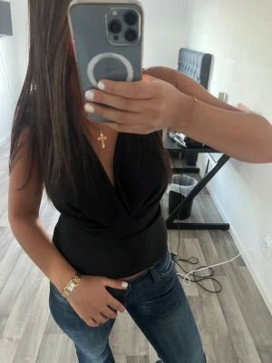 Svart omlott tubtopp - Snygg svart tubtopp med omlott-detalj och djup v-ringning. Toppen är ärmlös och sitter tight mot kroppen, perfekt för en trendig och stilren look. Passar till både jeans och kjol för en cool outfit.