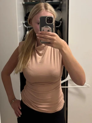 Beige ärmlös topp med axelvadd - Snygg beige topp med draperad halsringning och figurnära passform. Toppen är ärmlös och har en mjuk, stretchig känsla som gör den bekväm att bära. Perfekt att styla med både jeans och kjol för en trendig look.