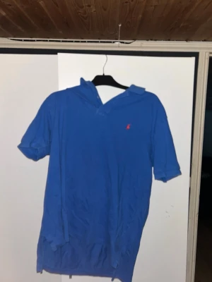 Blå pikétröja från Ralph Lauren - Klassisk blå pikétröja från Ralph Lauren med korta ärmar och krage. Tröjan har den ikoniska röda broderade loggan på bröstet. Tillverkad i mjuk bomull som känns skön mot huden. Perfekt för en avslappnad och stilren look.