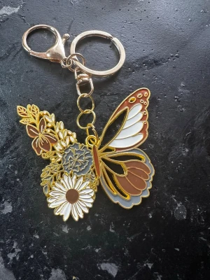 Väsksmycke/Nyckelring fjäril och blommor guld - Snygg väsksmycke /nyckelring i metall med fjäril och blommor i vitt, brunt, grått och guld. Detaljerad design med emaljfärger och guldfärgade kanter. Perfekt att hänga på väskan eller nyckelknippan för en personlig touch.