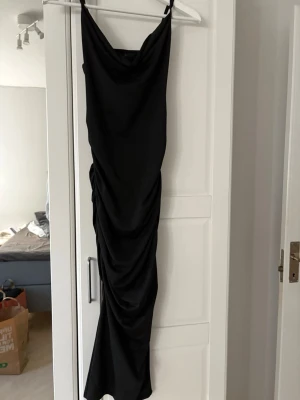 Svart lång klänning från SHEIN - Svart lång klänning från SHEIN med smala axelband och draperad design längs sidorna. Klänningen har en figurnära passform och är tillverkad i ett mjukt, stretchigt material som ger en snygg siluett.
