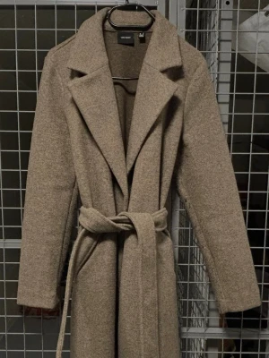 Mörk Beige kappa  - Stilren beige kappa med breda slag och knytskärp i midjan. Kappan har en rak passform och är tillverkad i ett mjukt, ull-liknande material. Perfekt för dig som vill ha en klassisk och trendig look under kyliga dagar.
