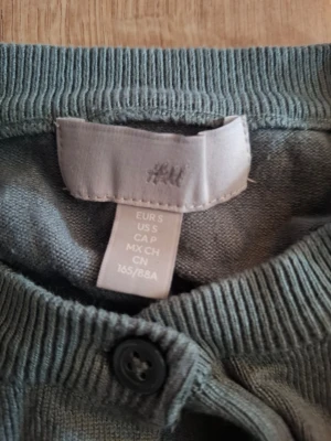 Grön stickad tröja från H&M - En enkel och stilren grå stickad tröja från H&M i storlek S. Tröjan har rund halsringning, ribbade muddar vid ärmslut och nederkant samt en knappdetalj vid halsen. Perfekt för lager på lager och passar till det mesta.